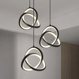 Three modern pendant lights with intertwined, glowing LED rings hang from the ceiling in a minimalist room with white walls and a wooden accent.
