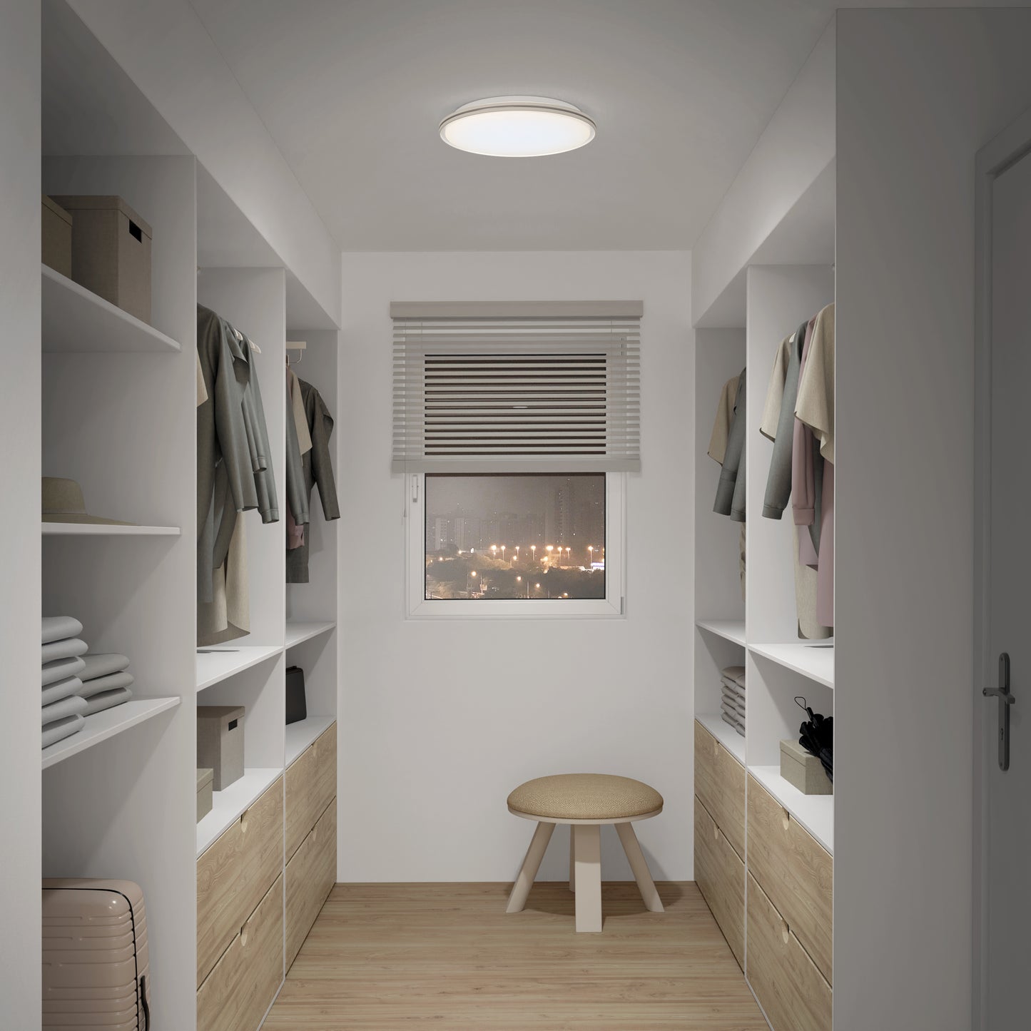 A modern walk-in closet features shelves and drawers on both sides, neatly hung clothes, a round stool by a window with blinds, and is brightly lit by an Artika Astrid 16.5 in. LED flush mount light with remote.