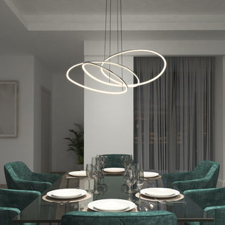 A modern dining room with a glass table for six, teal velvet chairs, and the Artika Swirl Butterfly LED contemporary pendant light (3 CCT), with large curtains and a potted plant in the background.