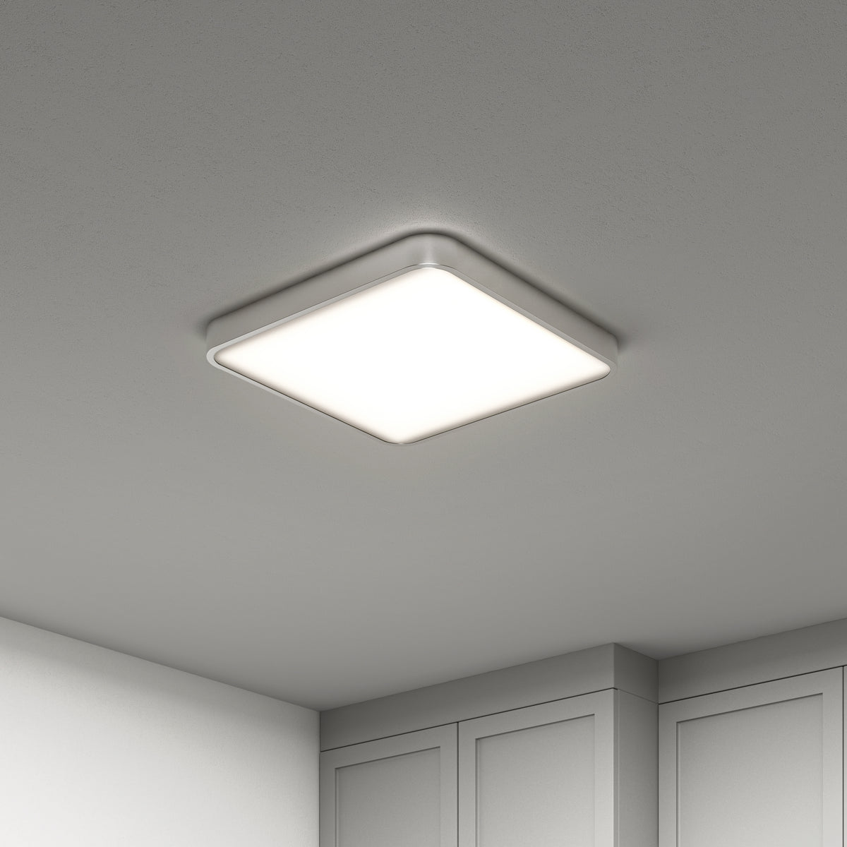 A modern, square-shaped ceiling light fixture with rounded edges emits a bright, even glow in a minimalist room with white walls and cabinets.