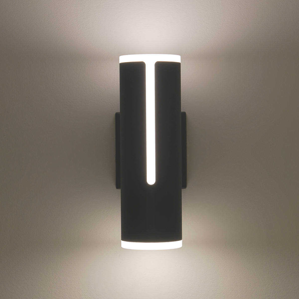 A modern black wall sconce with cylindrical shape emits soft white light from the top, bottom, and a vertical slit in the center, casting a gentle glow on the wall.
