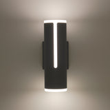 A modern black wall sconce with cylindrical shape emits soft white light from the top, bottom, and a vertical slit in the center, casting a gentle glow on the wall.