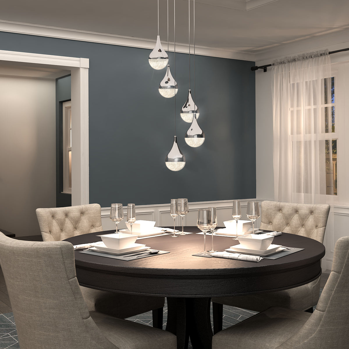 A modern dining room with a round dark wood table set for four, beige upholstered chairs, blue-gray walls, large window with sheer curtains, and five teardrop pendant lights hanging above the table.