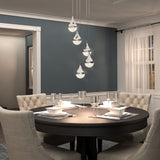 A modern dining room with a round dark wood table set for four, beige upholstered chairs, blue-gray walls, large window with sheer curtains, and five teardrop pendant lights hanging above the table.