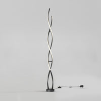 The Artika Swirl modern LED floor lamp features two black intertwining curved stems, one emitting soft white light. It stands on a round base with a visible plug-in cord, set against a plain light gray background.