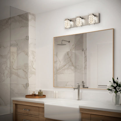 Modern bathroom with a marble shower, large mirror, wooden vanity, white countertop, chrome faucet, and a vase of white flowers. Three-light fixture with crystal details is mounted above the mirror.