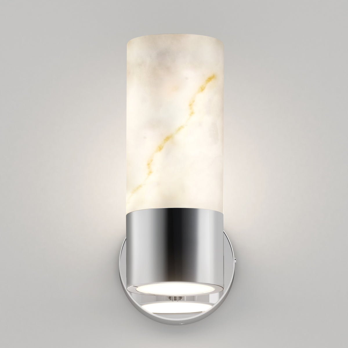 A modern wall sconce with a cylindrical white alabaster shade featuring light brown veining, mounted on a shiny silver metal base, emits a soft, diffused light against a plain gray wall.