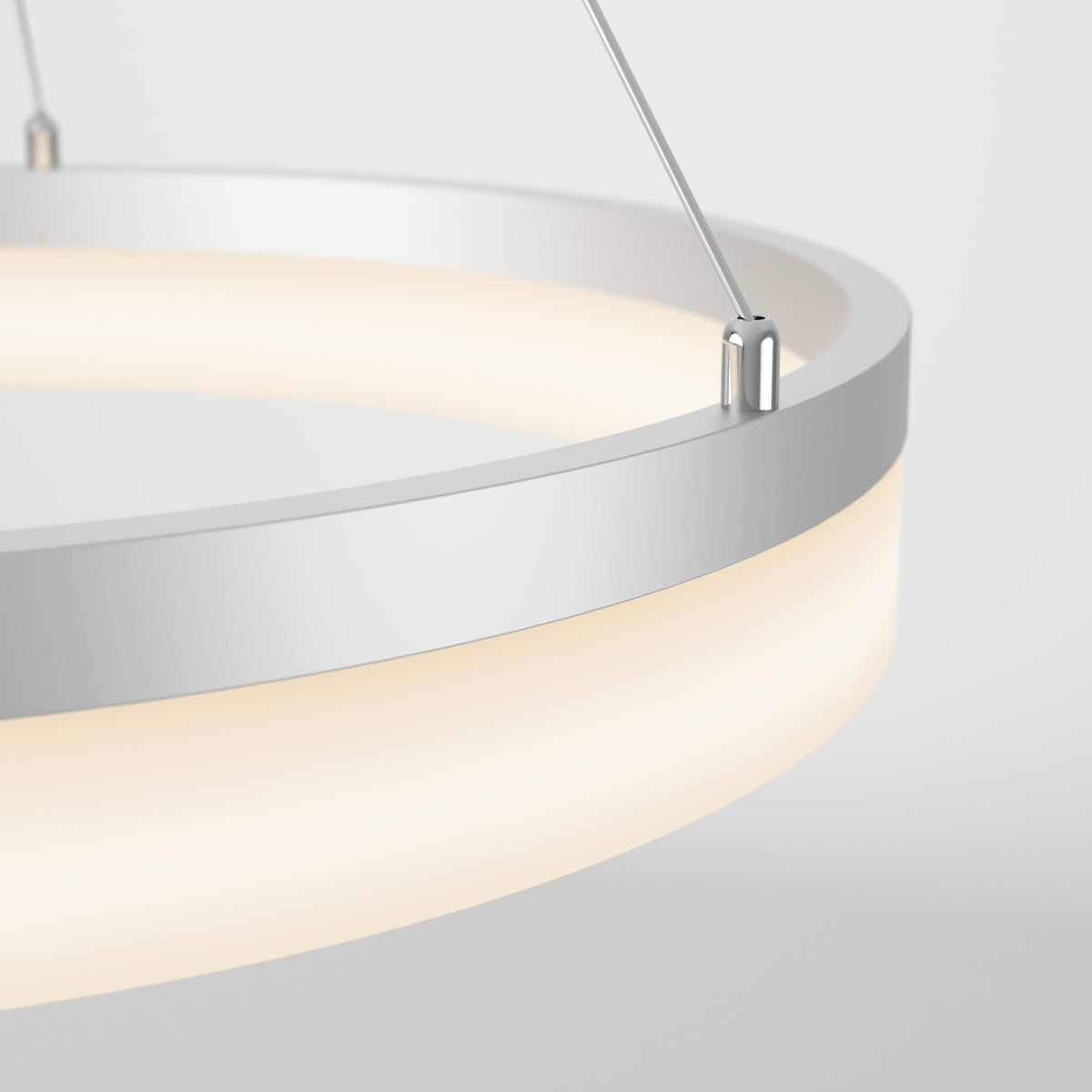 A close-up of a modern circular pendant light with a white and silver frame, emitting a soft, warm glow. The fixture is suspended by thin metal wires against a plain light background.