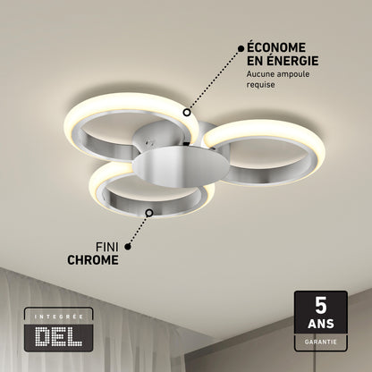 The Artika Nyala 15 in. LED flush mount light 5CCT features three circular chrome-finished LED rings mounted on a white ceiling. French text notes its energy efficiency and 5-year warranty. A curtain appears partly in the bottom left corner.