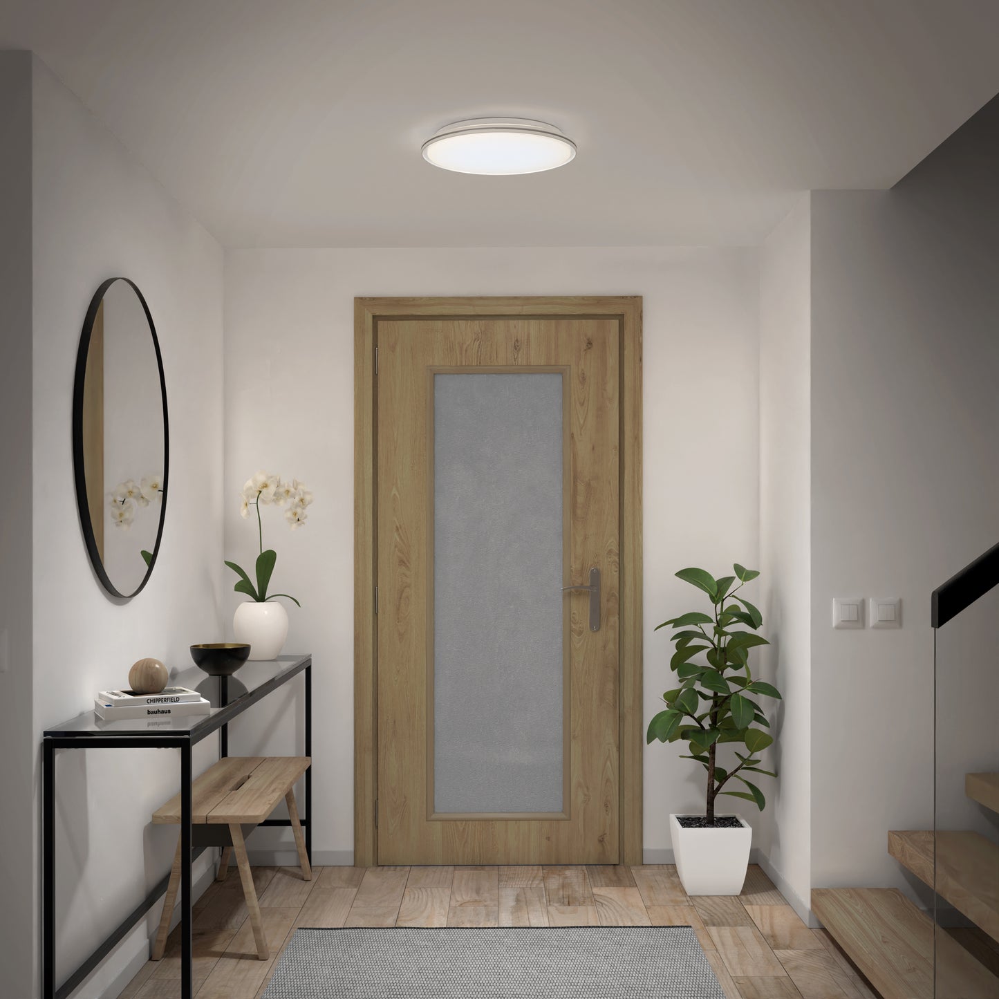 A modern hallway features a wooden door, console table with decor, round mirror, bench, potted plants, and staircase. The space is brightened by the Artika Astrid 16.5 in. LED flush mount light with 5 CCT and remote, enhancing its minimalist decor.