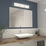A modern bathroom with white subway tiles, a large rectangular mirror, a wooden countertop with a white vessel sink, a silver faucet, a potted plant, toiletries, and a rectangular light fixture above the mirror.