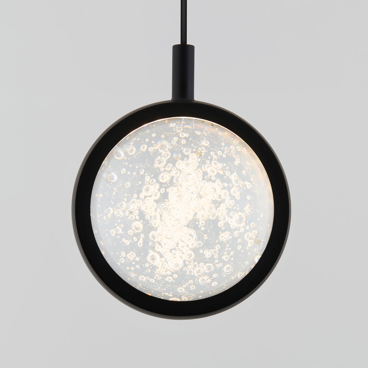A modern pendant light with a circular, black frame and a glass center featuring bubble-like patterns, suspended by a black cord against a plain, light gray background.