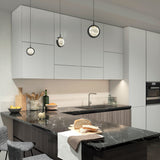 A modern kitchen with white cabinets, a black marble island, and hanging pendant lights. The island has a loaf of bread on a cutting board, a jar of pasta, and a white cloth. Cooking utensils and bottles are by the sink.