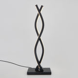 A modern table lamp with two twisting, black metal light strips on a square base, emitting a soft white light. The lamp is plugged in with a visible black cord against a plain light background.
