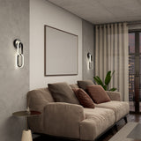 A modern living room with a beige sofa and assorted cushions, a small round side table, wall-mounted lights, a large framed artwork, indoor plants, and floor-to-ceiling curtains by a glass door.