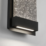A modern wall-mounted light fixture with a black frame and a clear panel filled with small, sparkling bubbles, emitting a soft white light onto a light gray wall.