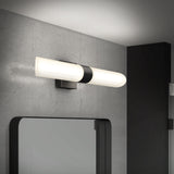 Modern wall-mounted cylindrical light fixture with a black base, installed above a large mirror on a gray concrete wall in a contemporary bathroom setting.