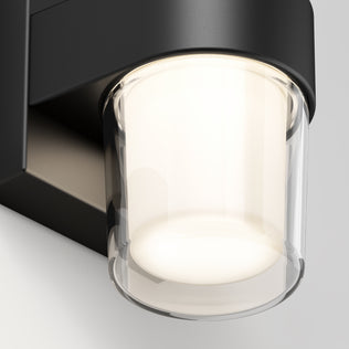 A close-up of the Artika Halden indoor-outdoor LED wall light 5 CCT showcases its cylindrical glass shade emitting a soft, warm glow against a light-colored wall.