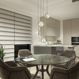 A modern kitchen and dining area with neutral tones, a round glass table with four padded chairs, pendant lights above, a potted plant, and horizontal striped blinds on a large window.