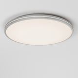 A modern, round ceiling light with a white diffuser and a silver or gray trim, mounted on a plain white ceiling, emitting a soft, even glow.
