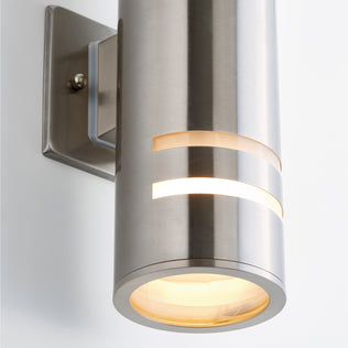 Close-up of the Artika Voltz indoor-outdoor wall light, a modern brushed metal cylinder with a frosted lens, emitting warm light through horizontal slits and an open bottom, mounted on a rectangular metal plate.