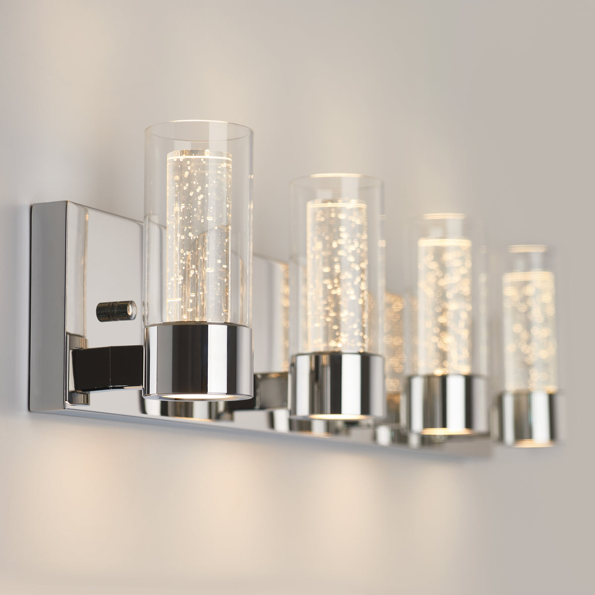 A modern wall-mounted light fixture with four cylindrical glass shades, each containing LED bubbles, and a sleek chrome base, illuminating a neutral-colored wall.