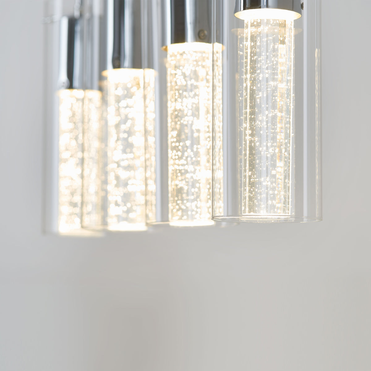 Close-up of modern cylindrical pendant lights with clear glass and bubbles inside, emitting a bright white glow against a plain light background.