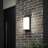 A modern rectangular wall light with a frosted cover is mounted on a brick exterior wall next to a dark-colored door, with green foliage visible at the bottom right corner.