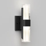 A modern wall sconce with a rectangular black metal base and a vertical frosted glass light fixture, emitting a soft white glow against a plain light gray wall.
