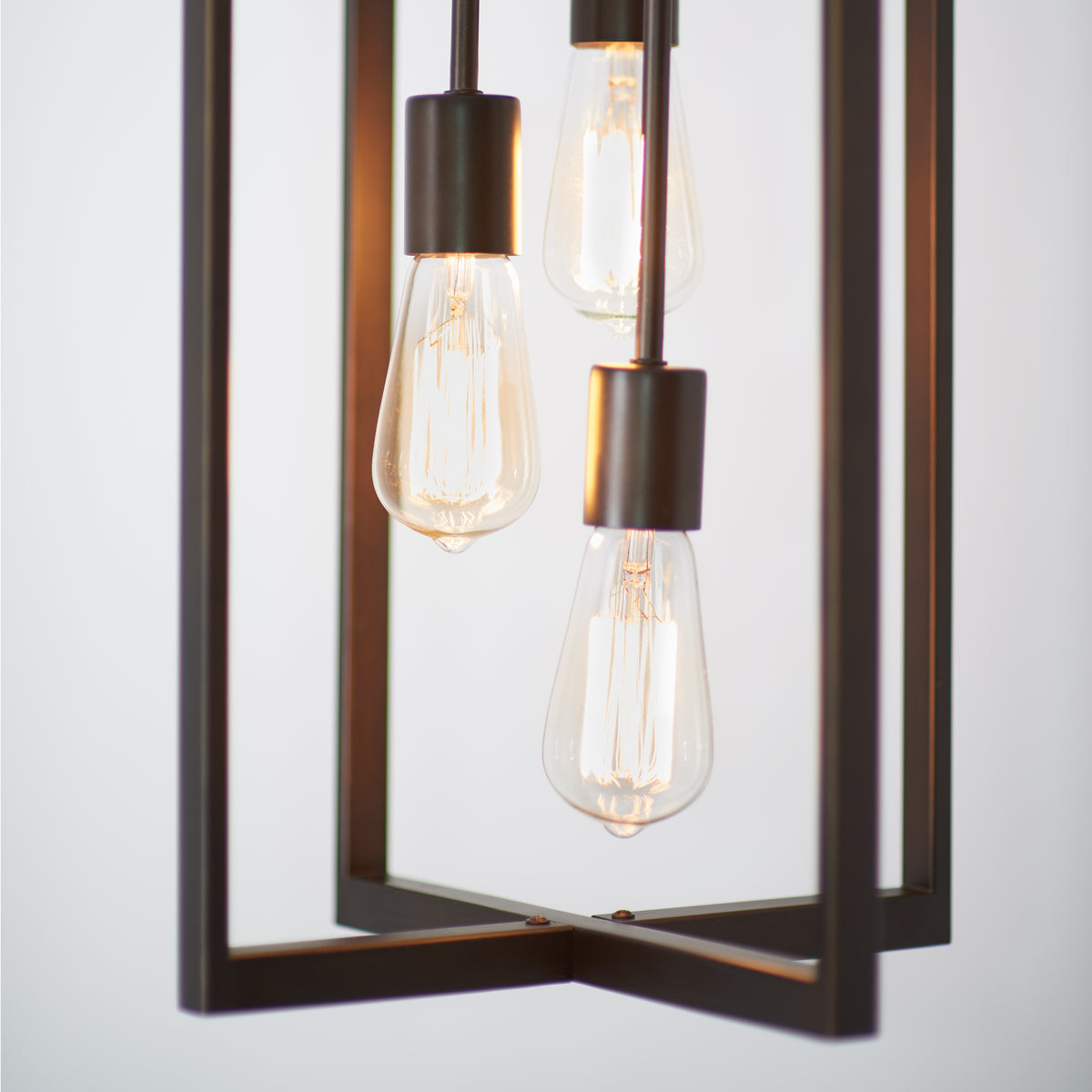 Close-up of a modern light fixture with three exposed Edison-style bulbs suspended inside a rectangular metal frame. The bulbs emit a warm glow against a plain, light background.