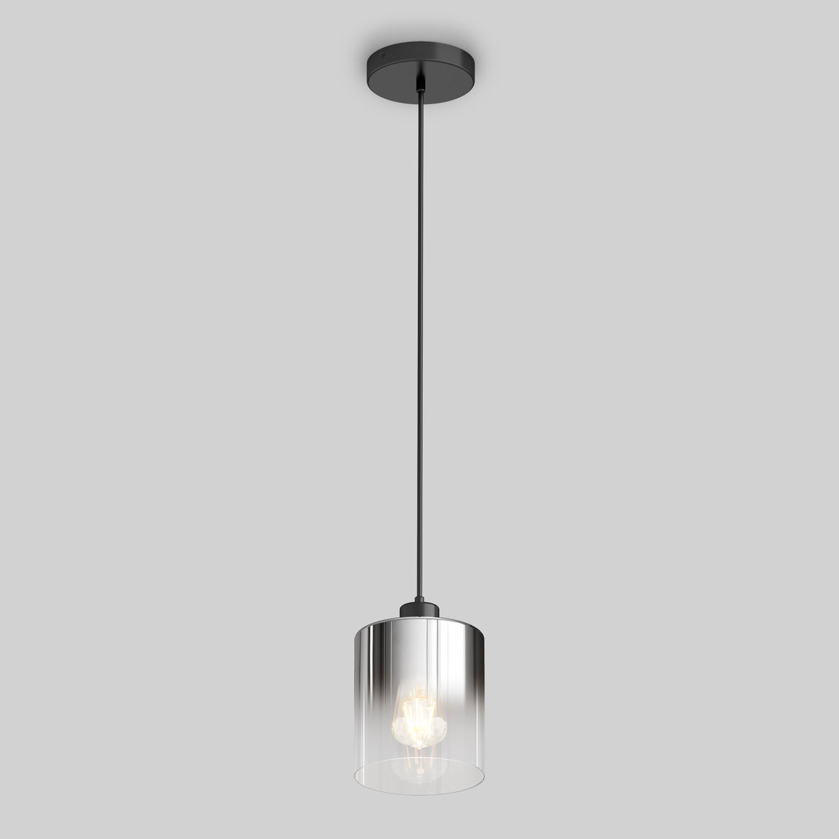 A modern pendant light with a cylindrical clear glass shade hangs from a black cord and ceiling mount against a plain light gray background. The bulb inside the shade is illuminated.