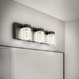 A modern bathroom wall light fixture with three square, crystal-like glass shades mounted on a rectangular black base, installed above a large mirror on a light gray wall.