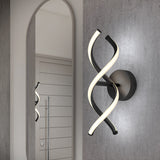 A modern wall sconce with two intertwined, curved LED light bars mounted on a round black base, installed on a gray wall next to an arched mirror reflecting part of the fixture.