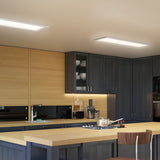 A modern kitchen features dark cabinets, light wood countertops, built-in appliances, and a Sunray ultra thin LED light panel by Artika. Dishes are visible in a glass-front cabinet, with a faucet, blender, utensils on the counter, and a wooden chair in front.