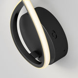 Close-up of a modern black wall-mounted LED light fixture with a circular base and a curved, illuminated strip, set against a plain light gray background.