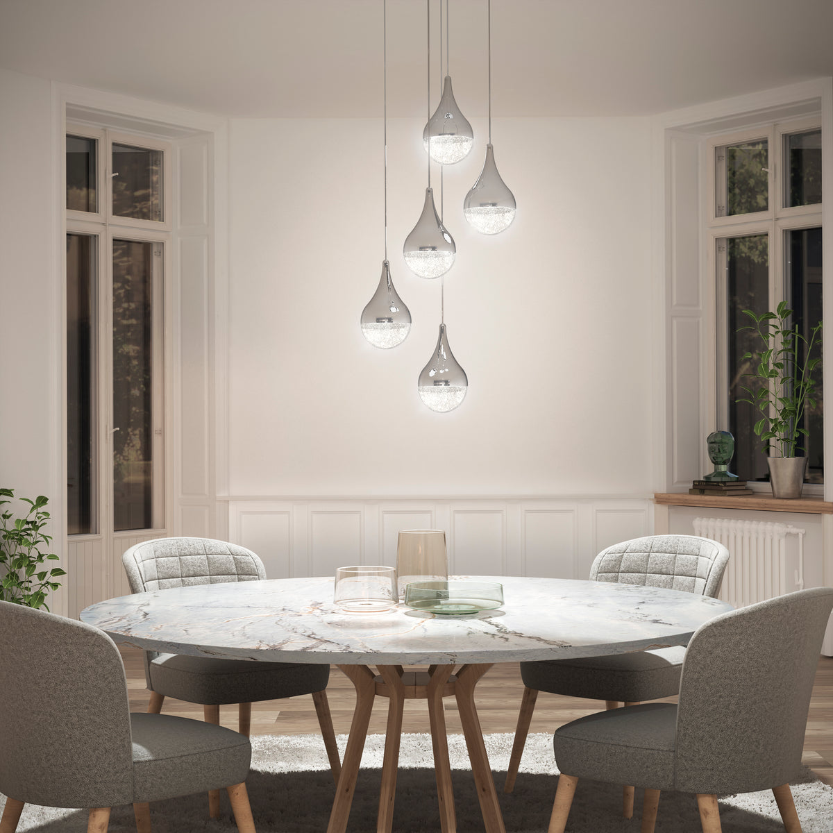 A dining room with a round marble table, four upholstered gray chairs, and five modern glass pendant lights hanging above. Large windows let in natural light; plants and decor sit on windowsills and the floor.