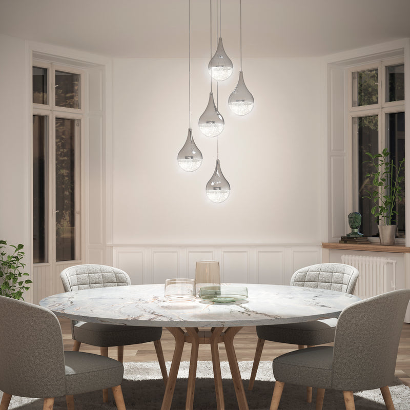 A bright, elegant dining area features a round marble table, four upholstered chairs, and the Artika Glitzer 5-light LED pendant hanging above large windows with minimal decor.
