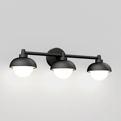 The Artika Boden LED mid-century bathroom vanity light features a modern black design with three dome shades and glowing round bulbs mounted on a horizontal bar, shown against a plain light gray wall.
