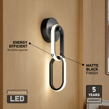 Modern wall sconce with a matte black finish and integrated LED, featuring two interlocking oval rings. Text highlights energy efficiency, no bulbs needed, and a 5-year warranty. Mounted on a light wood wall.