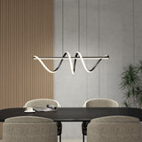A modern dining room with a black table, four beige chairs, a rolled-up magazine, a cup and saucer, and a sculptural wave-shaped pendant light hanging above. A plant and ribbed wooden wall accent are in the background.