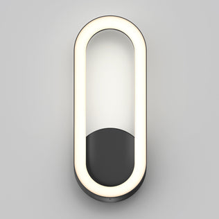 The Artika Aiden modern LED indoor-outdoor wall light 5 CCT features an oval LED with a black half-oval base, mounted on a light gray wall and emitting a soft, even glow.