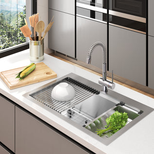 The Artika Newport 33 in. single bowl stainless steel workstation sink features a flexible faucet, a strainer with leafy greens, a white bowl on the drying rack, and a cutting board with corn and utensils by the window.