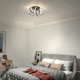 A modern bedroom with a gray cushioned bed, patterned bedding, decorative pillows, a small side table, a wooden desk with a chair, a wall mirror, and a unique, sculptural ceiling light fixture.