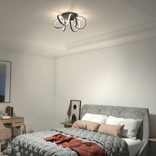A modern bedroom features a gray upholstered bed with a checkered blanket and pillows. The Swirl Ribbon flush mount light by Artika is on. A wooden desk with a mirror and chair sits near the bed.