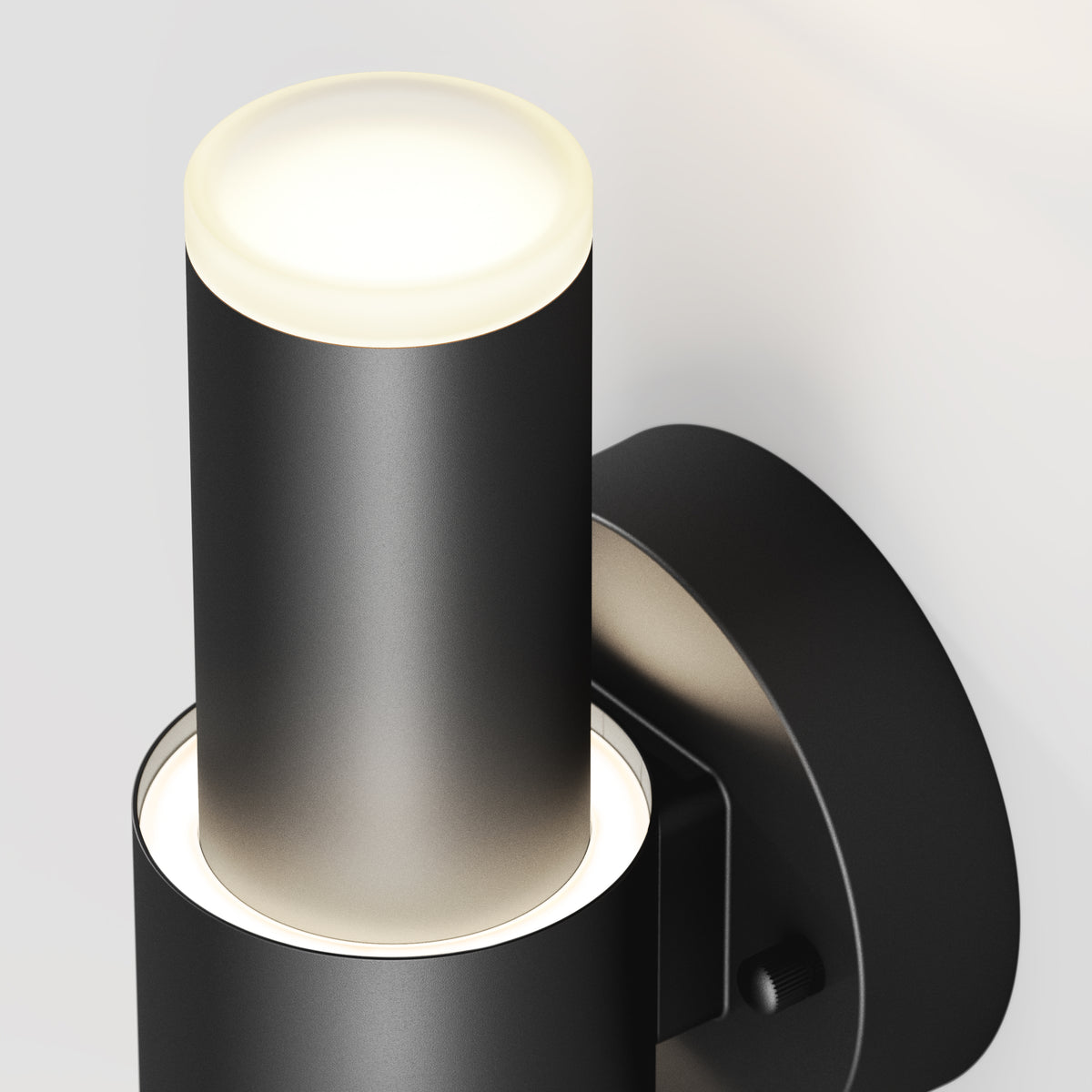 Close-up of a modern black cylindrical wall sconce with a frosted glowing top and a circular base, mounted on a light-colored wall. The light appears sleek and minimalist in design.