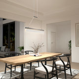 A modern dining room with a wooden table, six black chairs, a glass vase with branches, a linear pendant light above the table, large windows, and neutral-toned walls and decor.