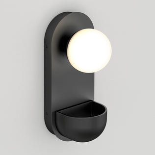 The Artika Bloomfield LED modern mid-century wall sconce 5 CCT features a matte black finish, a round frosted glass globe on top, and a semi-circular storage compartment, shown mounted on a pale gray wall.