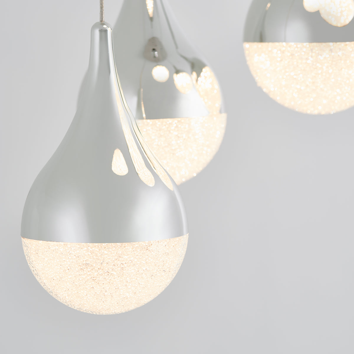Close-up of three modern pendant lights with a teardrop shape, silver tops, and textured frosted bottoms emitting a warm glow, set against a light gray background.