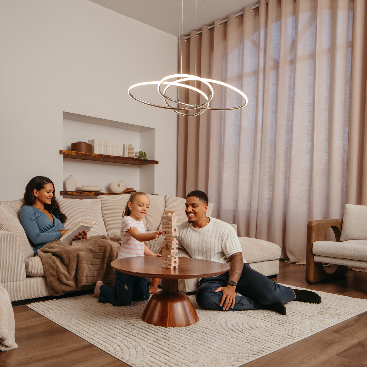 A family enjoys a warmly decorated, neutral-toned living room illuminated by the Artika Swirl Butterfly LED contemporary pendant light (3 CCT) as they relax and play together around a round coffee table.