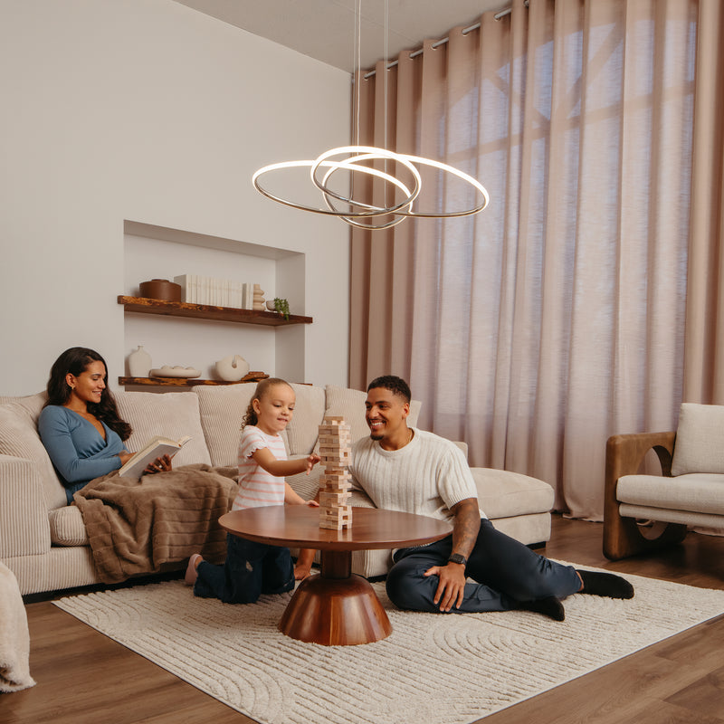 A family enjoys a warmly decorated, neutral-toned living room illuminated by the Artika Swirl Butterfly LED contemporary pendant light (3 CCT) as they relax and play together around a round coffee table.